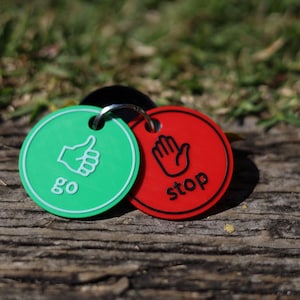May include: Two circular plastic tags connected by a metal ring. The green tag features a white thumbs-up icon and the word go. The red tag features a black open-hand icon and the word stop. Both tags have a raised border and flat surfaces.