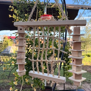 May include: A hanging bird toy featuring a central square rope net suspended between two horizontal wooden bars. Two vertical columns of alternating wooden blocks and flat wooden squares are attached to the sides, all connected by thick jute rope.