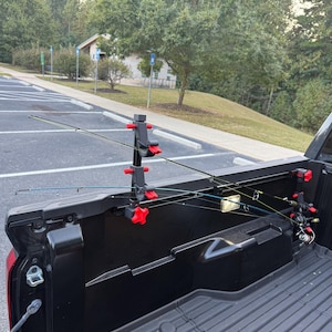 May include: A black truck bed fishing rod rack system featuring adjustable vertical mounts with bright red knobs and clamps, securely holding two fishing rods in place along the side rail of a black pickup truck bed.