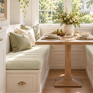 May include: A light green textured bench cushion with matching throw pillows sits on a white built-in storage bench. A light oak pedestal table is set with white ceramic bowls, plates, a wooden fruit bowl with pears, and a white vase with flowers.