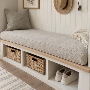 May include: A white entryway bench with a light gray textured cushion, a matching gray throw pillow, and a cream-colored knit throw blanket. Two woven wicker storage baskets sit in the lower cubbies, alongside a pair of white sneakers.