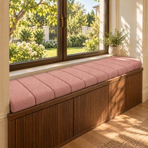 May include: A long, rectangular window seat featuring a plush, dusty pink upholstered cushion with vertical tufted detailing. The base is constructed from dark brown wood with a vertical slat design, positioned directly beneath a large window.