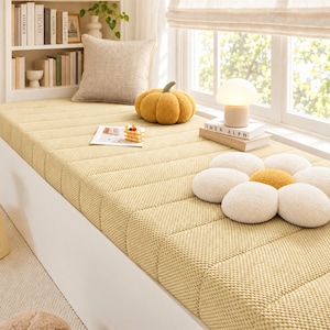 May include: A yellow textured bench cushion with a grid pattern, featuring a beige square pillow, a yellow pumpkin-shaped pillow, a white flower-shaped pillow, a small book, a miniature cake model, and a mushroom-shaped lamp on a book.