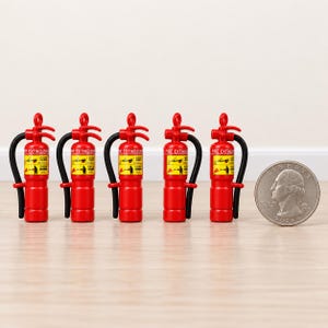 Peut inclure: Cinq extincteurs miniatures rouges avec des tuyaux noirs et des étiquettes jaunes portant l'inscription FIRE EXTINGUISHER, présentés à côté d'une pièce de monnaie pour donner une échelle de la petite taille de ces objets décoratifs.