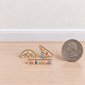 Puede incluir: Tres herramientas de dibujo en miniatura de madera que incluyen un transportador, una escuadra y una regla recta, cada una decorada con coloridos patrones florales y motivos de costura. Una moneda de cuarto de dólar sirve de referencia.