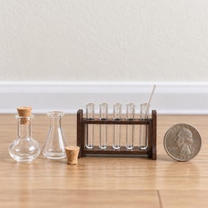 May include: A miniature laboratory set featuring two clear glass bottles with cork stoppers, a dark brown wooden test tube rack holding five clear glass test tubes, and one glass stirring rod, displayed next to a standard US quarter coin.