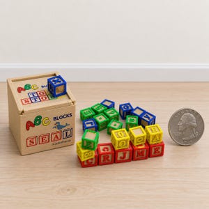Può includere: Una scatola portaoggetti in miniatura in legno con l'etichetta ABC BLOCKS SEAL e un blocco blu sopra, accompagnata da una collezione di piccoli blocchi alfabetici colorati in rosso, verde, blu e giallo, accanto a una moneta da 25 centesimi USA.