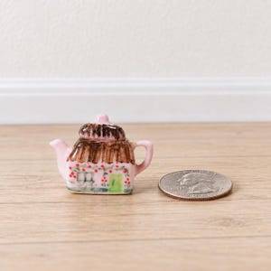 May include: A miniature ceramic teapot shaped like a cottage with a brown thatched roof, pink walls, a green door, and red floral accents. The teapot is positioned next to a United States quarter coin for scale, showing its small size.