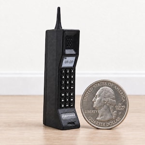 May include: A miniature black Motorola brick mobile phone featuring a small screen showing 08:00, a numeric keypad, and a top antenna. The device is placed beside a United States quarter coin, which provides a clear sense of its small scale in centimetres.