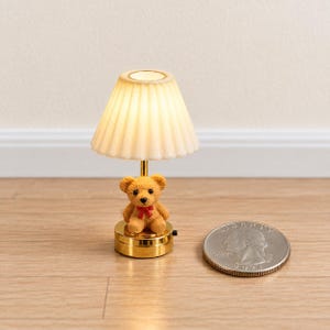 Può includere: Una lampada da tavolo in miniatura con un paralume plissettato color bianco sporco e una base dorata con un piccolo orsacchiotto marrone con un fiocco rosso. La lampada è posizionata accanto a una moneta statunitense per indicarne le dimensioni.