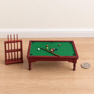 Pode incluir: Uma mesa de bilhar em miniatura de madeira com superfície de feltro verde, seis caçapas e pernas de madeira escura. O conjunto inclui um suporte de madeira com quatro tacos, um taco sobre a mesa e várias pequenas bolas de bilhar coloridas.