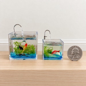 Op de afbeelding: Twee vierkante, doorzichtige harsbakjes gevuld met blauw water, groene waterplanten en kleine visfiguren. Elk bakje heeft een zilverkleurige kraanuitloop aan de bovenkant. Een kwartdollar munt ligt naast het kleinere bakje.