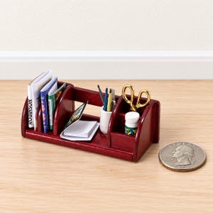 Op de afbeelding: Een miniatuur bureau-organizer van donker hout met kleine boeken, een witte pennenhouder met pennen, een gouden schaar en een klein flesje. Een munt van 25 cent ligt ernaast om de schaal van het miniatuurvoorwerp aan te geven.