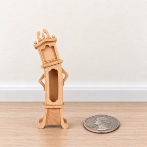 May include: A miniature light-colored wooden grandfather clock with a curved, leaning frame and an open face. The clock is positioned next to a United States quarter coin for scale, which has a diameter of approximately 0.955 inches.
