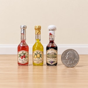 Op de afbeelding: Drie miniatuur glazen flesjes met labels waarop Berry Rosé, Golden Orchard en Vintage Cellar staat, met zilveren, gouden en witte folie omwikkelde doppen, geplaatst naast een Amerikaanse kwart dollar munt voor schaalvergelijking.