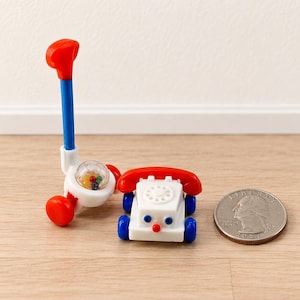 May include: A miniature toy push-along lawn mower with a blue handle and red wheels, and a miniature toy telephone on wheels with a white body, red receiver, and blue wheels, positioned next to a standard United States quarter coin for scale.