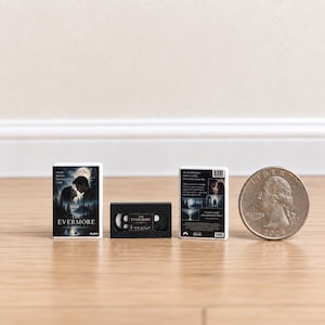 May include: A miniature VHS tape set titled The Evermore, including a front cover, a back cover, and a small black cassette tape. The items are displayed next to a United States quarter coin for scale, showing the small size of the movie memorabilia.