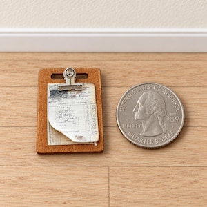 Könnte beinhalten: Ein Miniatur-Klemmbrett aus Holz mit einer silbernen Metallklemme, die ein kleines Stück Papier mit gedrucktem Text und Linien hält. Das Klemmbrett liegt neben einer amerikanischen Quarter-Münze, um die geringe Größe zu verdeutlichen.