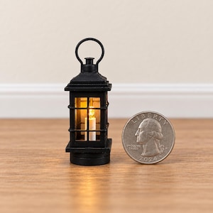 Pode incluir: Uma lanterna preta em miniatura com estrutura quadrada e alça superior, contendo uma pequena luz interna que brilha com um tom amarelo quente. Uma moeda de 25 centavos dos EUA está ao lado para demonstrar a pequena escala do objeto.