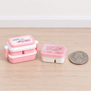 May include: A miniature two-tiered pink and white bento box with a cartoon bear illustration on the lid, next to a smaller rectangular pink and white container with a clear lid containing tiny utensils, positioned beside a US quarter coin.