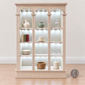 Könnte beinhalten: Ein heller Miniatur-Vitrinenschrank aus Holz mit drei bogenförmigen Abschnitten und integrierter LED-Beleuchtung. Die Regale enthalten kleine Dekorationsgegenstände wie einen Krug, eine Vase, eine Glasflasche, Bücher, eine Teetasse, Teller, Gläser, einen geflochtenen Korb und ein Gefäß. Eine Münze liegt daneben als Größenvergleich.