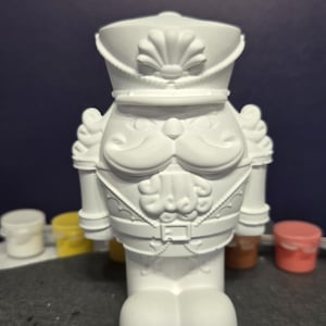 May include: A solid white, unpainted nutcracker figurine featuring a detailed military-style hat with a shell emblem, a prominent mustache, a jacket with epaulets, a belt with a buckle, and rounded feet, ready for decorative painting.