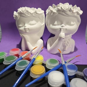 May include: Two white ceramic head planters featuring floral crowns, one with a hand on the cheek and one with a finger to the lips. Included are several small plastic pots of assorted acrylic paints and three blue-handled paintbrushes.