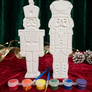 May include: Two white, unpainted nutcracker figurines made of a smooth material, standing upright. Below them is a plastic strip containing six small pots of paint in red, orange, yellow, green, blue, and purple, along with two thin paintbrushes.
