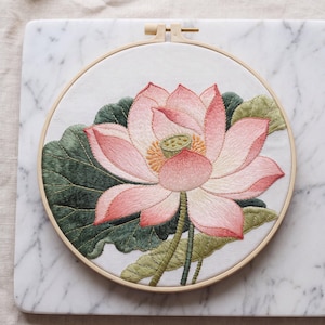 May include: A circular wooden embroidery hoop containing white fabric with a detailed needlepoint design of a large lotus flower. The flower features layered pink and white petals, a central green seed pod, and dark green textured leaves.