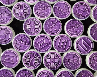 May include: A collection of round, purple wax seals featuring embossed designs related to the music group BTS. The designs include the BTS logo, the group's light stick, a finger heart gesture, and a circular border containing the members' names.