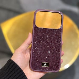 May include: A rectangular smartphone case featuring a deep purple, textured, crystal-encrusted back panel and a smooth, reflective gold-toned camera cutout area. A small, rectangular black plaque at the bottom displays the text The Bling World.
