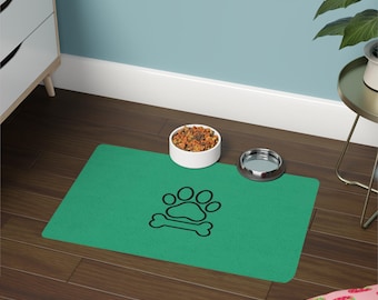 Può includere: Un tappetino rettangolare verde per l'alimentazione di animali domestici con un disegno a linee nere di un'impronta sopra un osso. Il tappetino è posizionato su un pavimento in legno scuro con una ciotola bianca piena di croccantini e una ciotola in metallo argentato vuota.