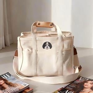 May include: A cream-colored canvas tote bag featuring a brown and white dog illustration on the front. The bag has two top handles with a tan leather grip, an adjustable shoulder strap, and multiple exterior pockets for storage.