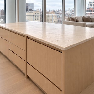 May include: A kitchen island featuring a light-toned wood base with flat-panel cabinetry and drawers. The rectangular countertop is made of a textured, cream-colored stone with a natural, porous surface finish and straight, clean edges.
