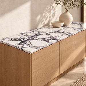 May include: A rectangular wooden sideboard featuring a polished white marble top with prominent dark purple and black veining. The cabinet has three flat-fronted light oak doors. A beige ceramic vase and a small matching bowl sit on the surface.