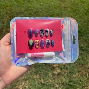 Puede incluir: Un juego de diez uñas postizas con diseños gráficos inspirados en anime en morado, rosa, amarillo y negro, presentadas sobre una tarjeta rosa dentro de una bolsa de plástico holográfica con un pequeño tubo de pegamento para su aplicación.