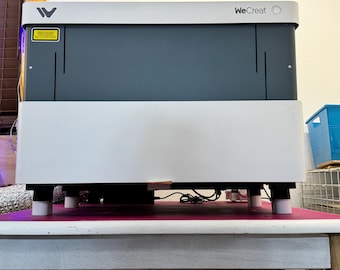 May include: A white laser engraver with the text "WeCreat" on the top. The engraver is on a white table with a pink surface underneath.