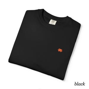 May include: A black crew neck t-shirt featuring a small, orange, pixelated character embroidered on the left chest. The shirt has a Comfort Colors brand label visible at the inner neckline, indicating a size large, and is folded neatly.