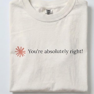 May include: An off-white crew neck t-shirt featuring a small, coral-coloured starburst graphic to the left of the black text that reads, "You're absolutely right!" The shirt has a Comfort Colors brand label visible at the inside of the neckline.