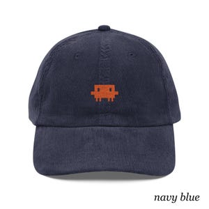 May include: A navy blue corduroy baseball cap featuring an embroidered orange pixelated alien character centered on the front panel. The cap has a structured crown, a curved brim, and a button at the top, with the text navy blue at the bottom.