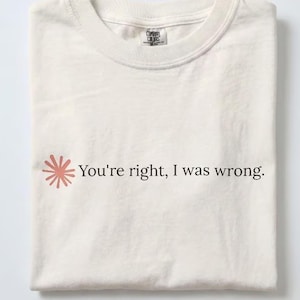 May include: An off-white crew neck t-shirt featuring a small, coral-coloured sunburst graphic to the left of the black text that reads, "You're right, I was wrong." The shirt has a visible brand label at the inner neckline and short sleeves.