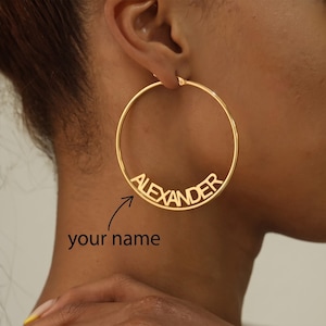 May include: A large gold-toned hoop earring featuring the name ALEXANDER cut out in capital letters across the center. Below the earring, a black arrow points to the text your name, indicating a customizable jewelry design.