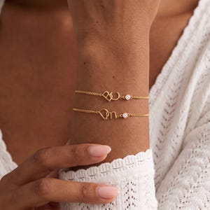 May include: Two gold-toned chain bracelets worn on the wrist. Each bracelet features a delicate metal heart charm connected to a lowercase letter, with a single small, round, clear stone set in a bezel mount positioned next to the letter charm.