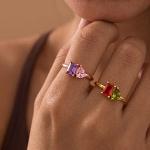 May include: Two gold-toned rings worn on fingers. One ring features a rectangular purple stone and a pear-shaped pink stone. The second ring features a rectangular red stone and a triangular green stone, both set in gold bands.
