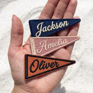 May include: Three triangular fabric pennant patches with embroidered names. The top patch is navy blue with the name Jackson in cream. The middle patch is dusty rose with the name Amelia in cream. The bottom patch is rust orange with the name Oliver in black.
