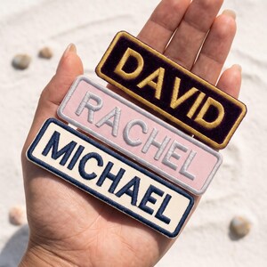 May include: Three rectangular embroidered name patches with stitched borders. The top patch is dark purple with gold text reading DAVID. The middle patch is light pink with silver text reading RACHEL. The bottom patch is cream with navy text reading MICHAEL.