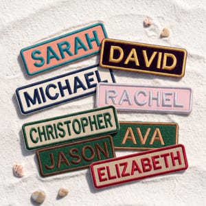 May include: A collection of rectangular embroidered name patches featuring the names SARAH, DAVID, MICHAEL, RACHEL, CHRISTOPHER, AVA, JASON, and ELIZABETH in various thread colors including teal, yellow, navy, white, green, and red on contrasting backgrounds.