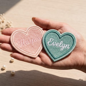 May include: Two heart-shaped embroidered patches with stitched borders. One patch is light pink with the name Harper embroidered in a matching thread. The other patch is sage green with the name Evelyn embroidered in white thread.