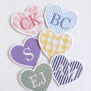 May include: Six heart-shaped fabric patches with white stitched borders. One features pink floral print with CK, one blue stripes with BC, one yellow gingham with M, one solid purple with S, one green polka dot with EJ, and one navy stripe with AM.