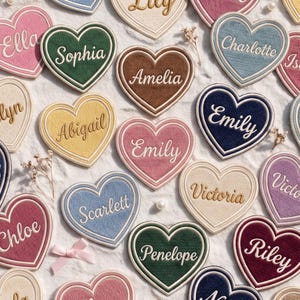May include: A collection of heart-shaped embroidered patches in various colors including pink, green, brown, blue, yellow, and cream. Each patch features a name stitched in white or gold thread, such as Sophia, Amelia, Emily, and Victoria.